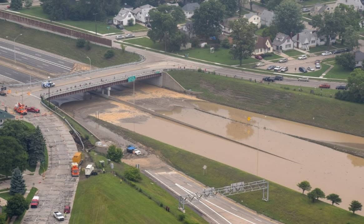 Aerial view of a flooded highway underpass in a residential area, with muddy water covering multiple lanes and emergency vehicles and construction crews responding nearby.