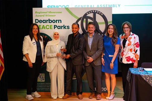 A group of six people stand smiling on stage as they accept the Regional Showcase Award for Dearborn PEACE Parks at SEMCOG’s General Assembly.