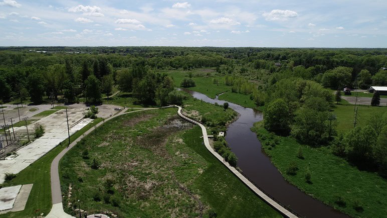 Aerial view of a winding river surrounded by lush greenery and wetlands, with a walking path and parking area nearby under a partly cloudy sky