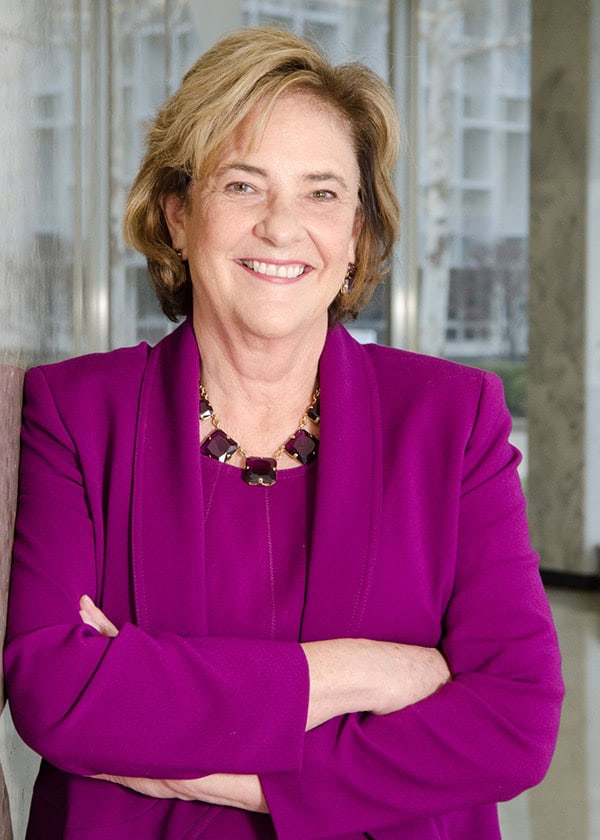 Gwen Markham, Chairperson at SEMCOG and Commissioner of Oakland County, smiling with arms crossed while wearing a bright purple blazer and necklace, standing in a modern indoor setting with glass and marble surroundings.