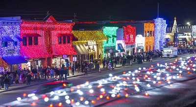 Downtown street at night decorated with colorful holiday lights, with crowds of people walking along lit-up storefronts.
