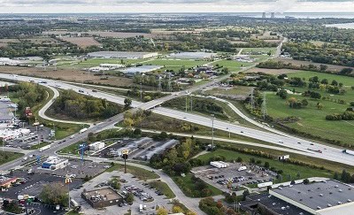 Aerial view of a highway interchange surrounded by commercial buildings, farmland, and green open space, with a river or lake visible in the distance