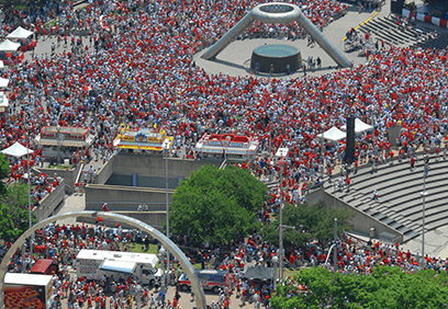 A large crowd of people, many wearing red, gathered in an urban plaza with food trucks, tents, and a central circular monument. The area includes trees, steps, and metal arch structures