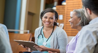 Smiling healthcare professional holding a clipboard and speaking with an older woman and others in a group setting