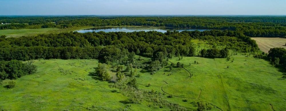 Aerial view of a green nature preserve with winding trails, dense forest, and a lake in the distance under a blue sky.