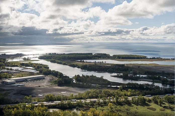 Aerial photo of a coastal area with wetlands, waterways, and industrial facilities near a large lake, under a cloudy sky with sunlight reflecting off the water.