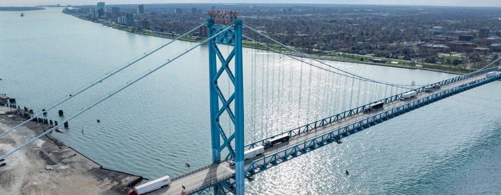 Aerial view of the Ambassador Bridge connecting Detroit, Michigan to Windsor, Ontario, with trucks crossing over the Detroit River on a sunny day.