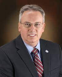 Portrait of the Chairperson of SEMCOG’s Finance and Budget Committee and Mayor of the City of Monroe. He is wearing a dark suit, striped red tie, glasses, and is smiling in front of a brown gradient background.