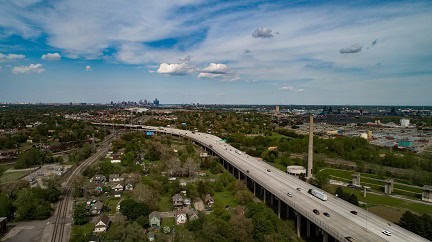 Aerial view of a highway overpass cutting through a residential and industrial area with the Detroit skyline in the distance under a partly cloudy sky