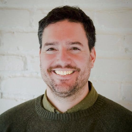 Christopher Girdwood, Co-Chairperson and CEO of the Detroit Region Aerotropolis Development, smiling in a casual professional headshot. He is wearing a green sweater over a collared shirt and standing in front of a white brick wall.