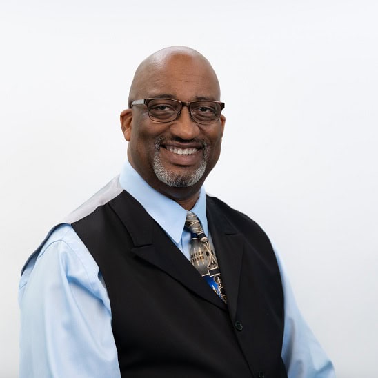 Donal Boynton, Co-Chairperson and Trustee of Van Buren Charter Township, smiling in a professional headshot. He is wearing glasses, a light blue shirt, patterned tie, and a black vest, posed in front of a white background.