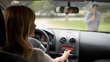 Woman driving car, distracted with changing radio station as a pedestrian in the background walks in front of the car