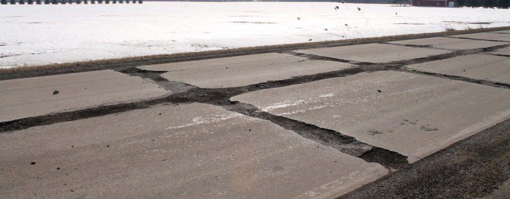 Cracked and deteriorating concrete road in a rural area with snow-covered fields, bare trees, and farm buildings in the distance.