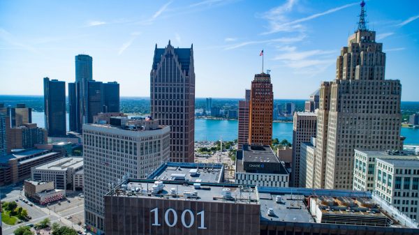 A daytime aerial view of downtown Detroit’s skyline with prominent high-rise buildings. The image shows the Renaissance Center towers on the left, several historic skyscrapers in the center and right, and the Detroit River in the background with Windsor, Ontario visible across the water. The foreground includes a building with “1001” displayed on its rooftop. The sky is clear with light streaks of clouds.