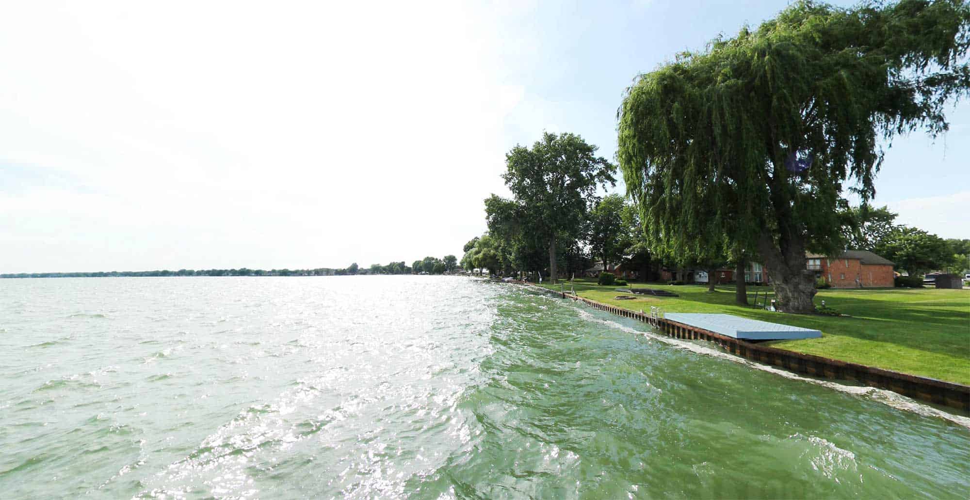 View of Lake St. Clair Water Trails showing a residential shoreline with a large willow tree, dock, and rippling green water under a bright sky. Homes and trees line the waterfront in the background.
