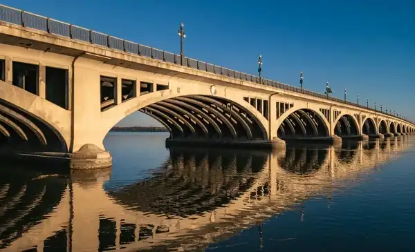 View of a large arched concrete bridge spanning over calm water, with strong reflections and a clear blue sky.