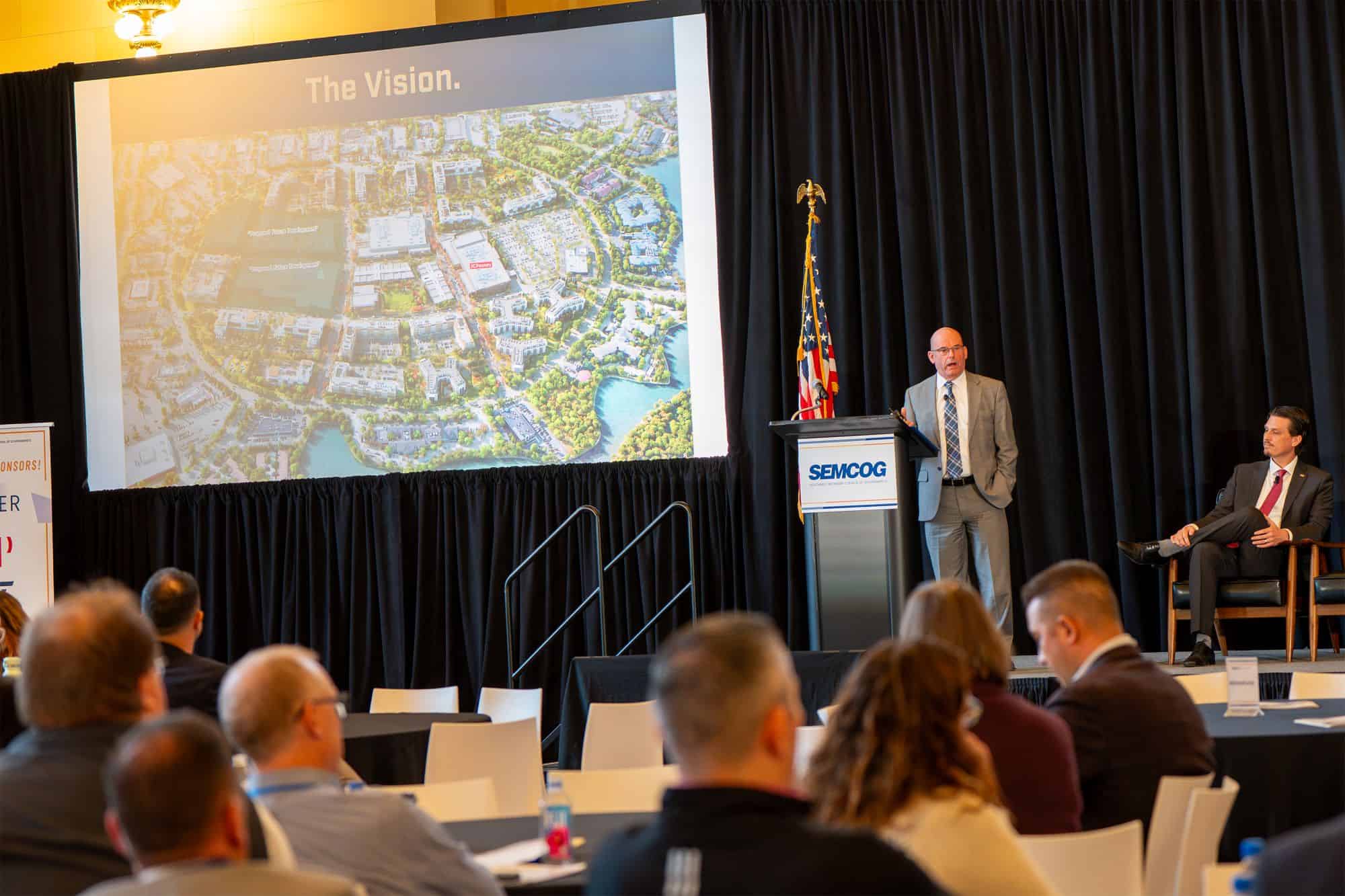 Mark Vanderpool speaking at a SEMCOG event, standing at a podium with the SEMCOG logo and American flags beside him. A large screen displays a presentation slide titled The Vision with an aerial development map. Audience members are seated and listening attentively.