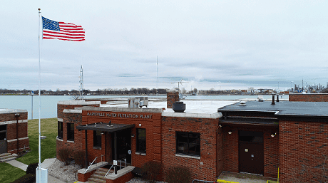 Exterior view of the Marysville Water Filtration Plant, a brick building situated along a waterfront with an American flag flying on a pole in the foreground and industrial structures visible across the water.