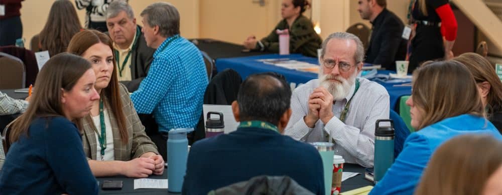 Members of the Oakland County Climate Task Force participate in a community meeting, seated around tables for group discussion on local climate action planning, with workshop materials and informational booths in the background.