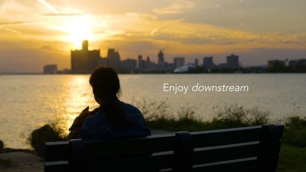 image of someone sitting on a bench with lake st Clair in the background while the sunsets over the Detroit skyline with the words 'Enjoy Downstream' written across the image