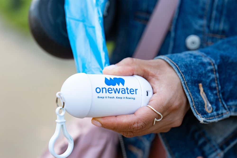 Person holding a white One Water-branded dog waste bag dispenser with blue bags, featuring the slogan Keep it fresh. Keep it flowing.
