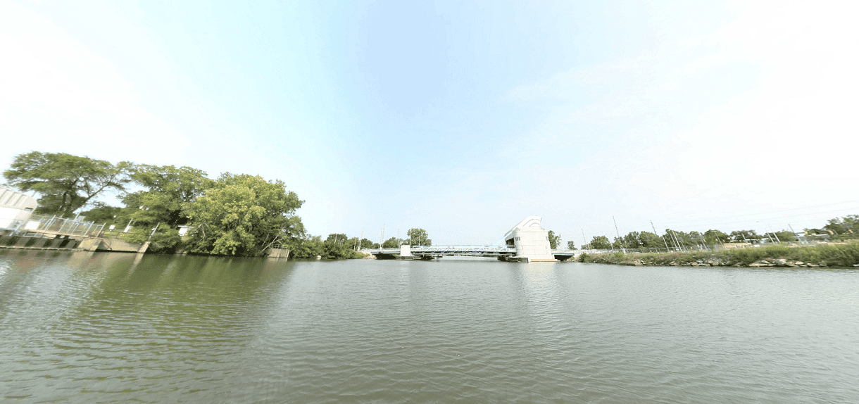 View of the Rouge River Water Trail (Industrial Route) featuring a drawbridge over calm water, with industrial fencing and trees on the left and a grassy shoreline with utility infrastructure on the right.