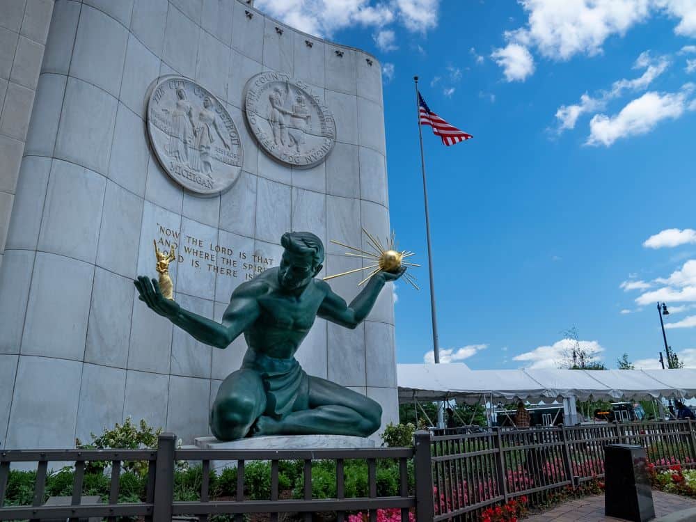 The Spirit of Detroit statue in front of the Coleman A. Young Municipal Center, with the American flag waving and blue sky overhead.