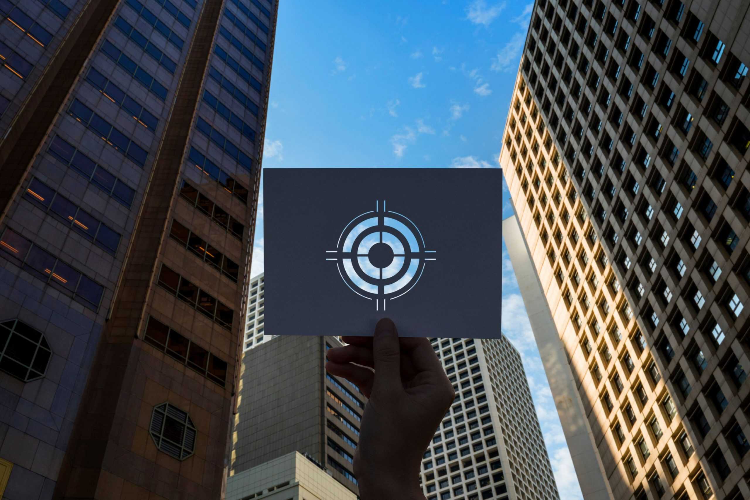 A hand holds up a card with a transparent target symbol cut out, aligning it with the blue sky and tall skyscrapers in the background, symbolizing vision, focus, or goal setting in an urban environment.