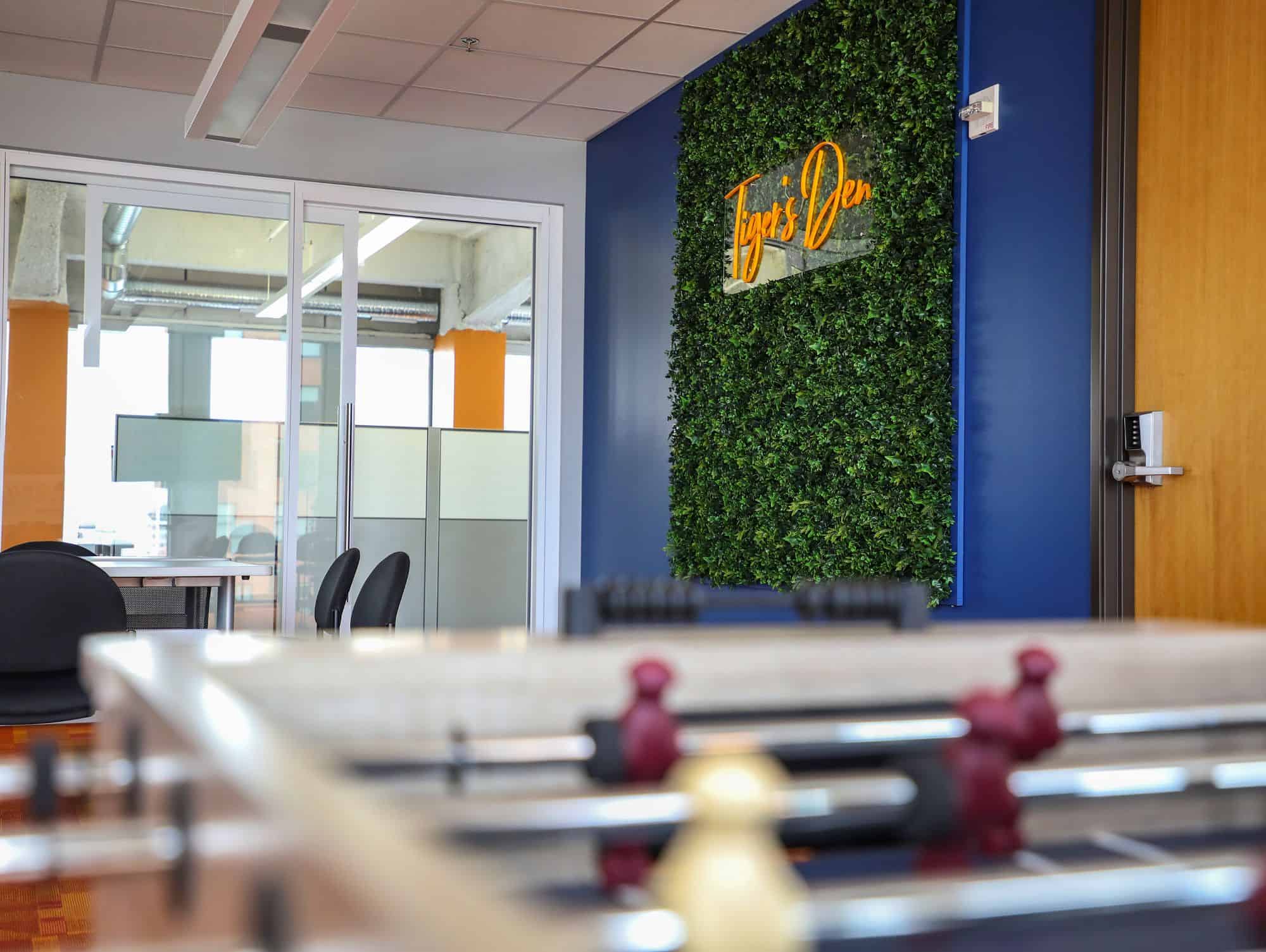 Modern office space featuring a foosball table in the foreground, a glass-walled meeting room with chairs in the background, and a vertical green wall with an orange neon sign that reads Tigers Den mounted on a dark blue accent wall.
