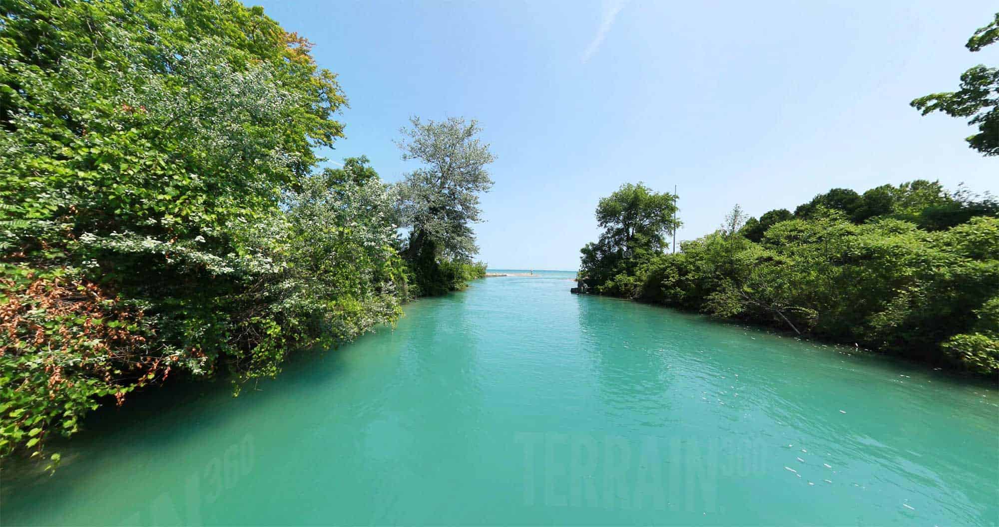 Clear turquoise river or channel flowing toward a large lake, bordered by dense green trees and vegetation under a sunny blue sky.