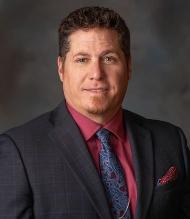 Robert McCraight, Vice Chair of SEMCOG’s Legislative Policy Platform Task Force and Mayor of the City of Romulus, posing for a professional headshot. He is wearing a dark plaid suit jacket, maroon shirt, and paisley tie with a coordinating pocket square, set against a dark gradient background.