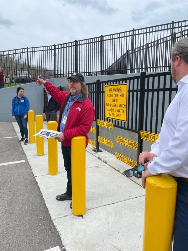 A person in a red jacket stands near yellow safety bollards and points toward an area beside a fenced flood‑control structure with multiple caution signs, while others observe.