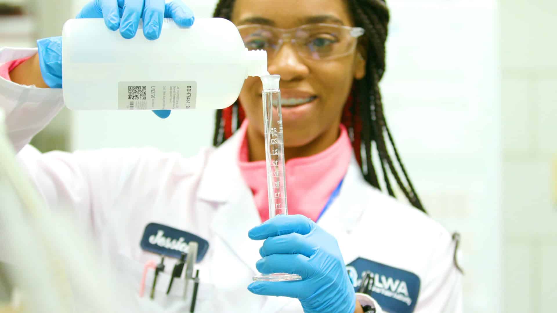 lab worker with gloves, safety glasses, and white coat, holding tube and pouring water, smiling