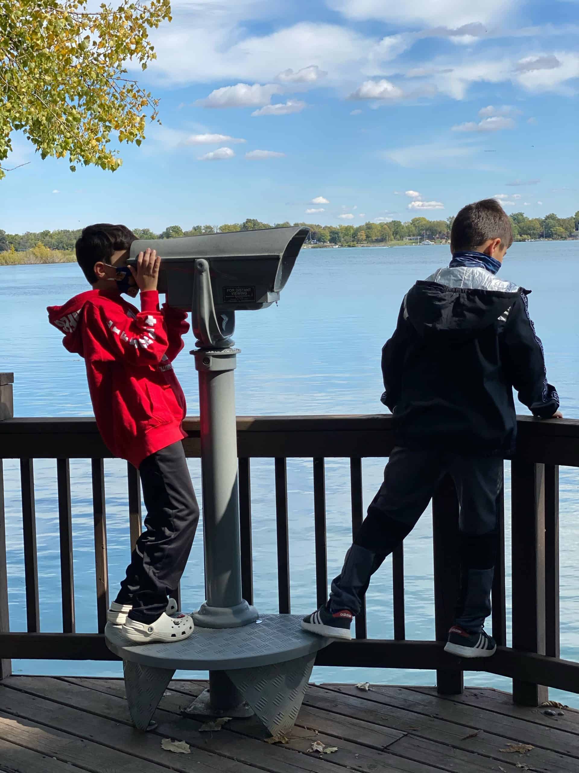 two young boys, one viewing the other with a telescope in front of a body of water