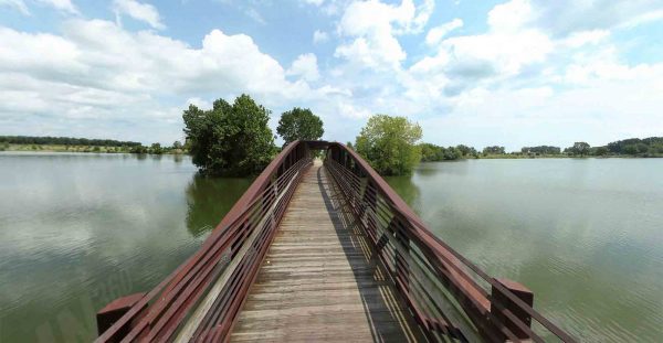 A wooden pedestrian bridge with red railings stretches over a calm lake toward a cluster of trees under a partly cloudy sky.
