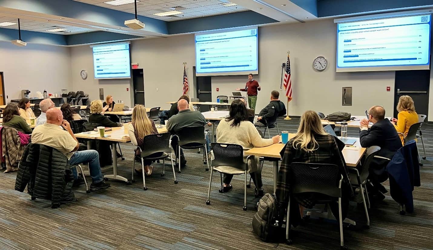 Meeting room with attendees sitting at round tables, speaker is at the front of the room with a presentation