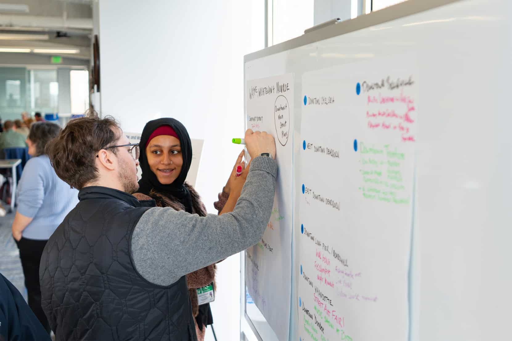 Someone writing with a marker on a collective idea board, someone is looking at them in a conversation