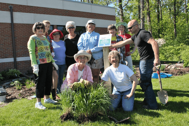 Group of people standing, wearing gardening clothes and gloves, holding shovels, next to rain garden sign