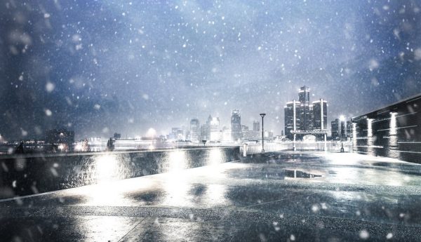 Snowy nighttime scene overlooking a wet waterfront promenade with bright lights reflecting on the ground and a distant city skyline in the background.