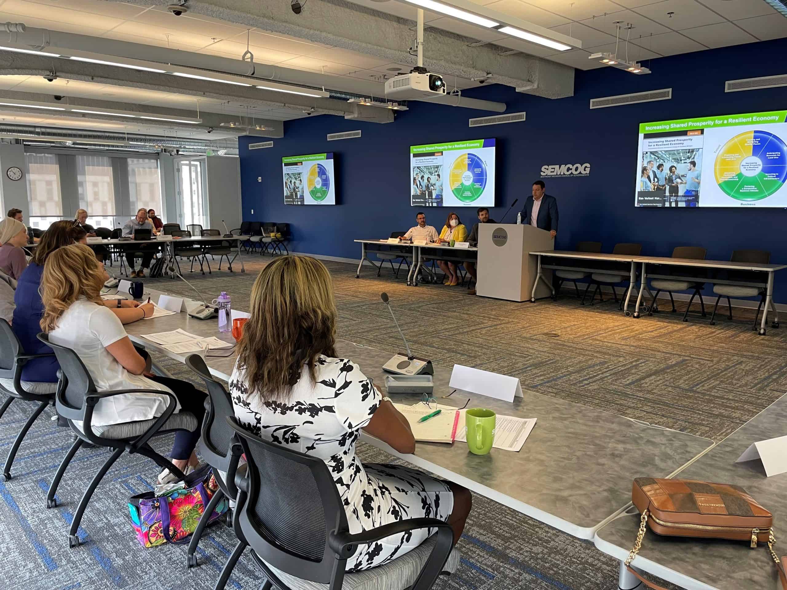 People seated around a conference table listening to a presenter at a podium, with presentation slides displayed on large screens in a meeting room.