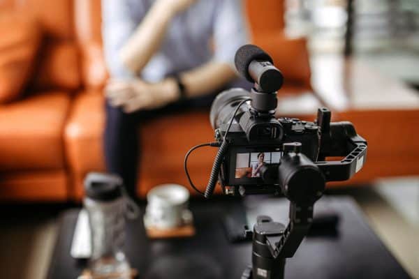 Video camera on a tripod recording a person seated on a couch in an indoor setting, with focus on the camera’s display screen.