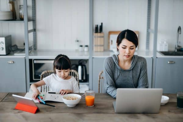 A person works on a laptop at a kitchen table while a child sits beside them drawing, with breakfast items and a tablet nearby.