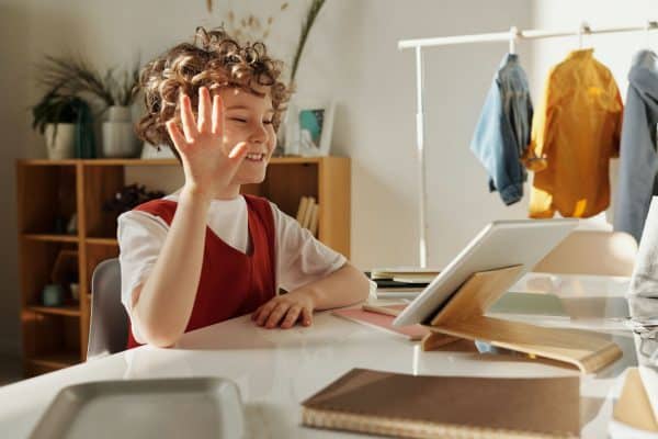 Child seated at a desk raising one hand while using a tablet on a stand, with shelves and hanging clothes in the background.