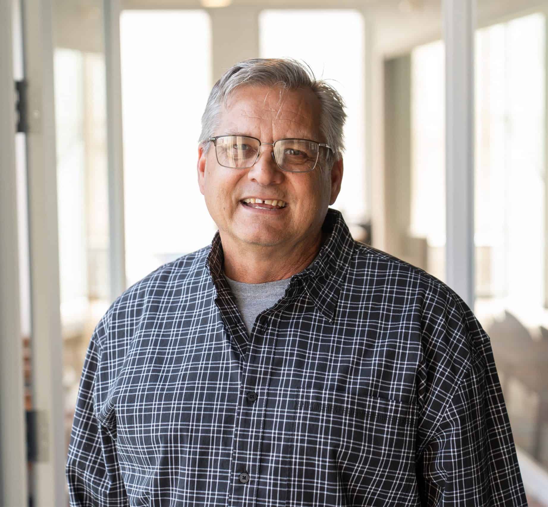 Portrait of a man named John, smiling with a blurred background of trees and buildings.