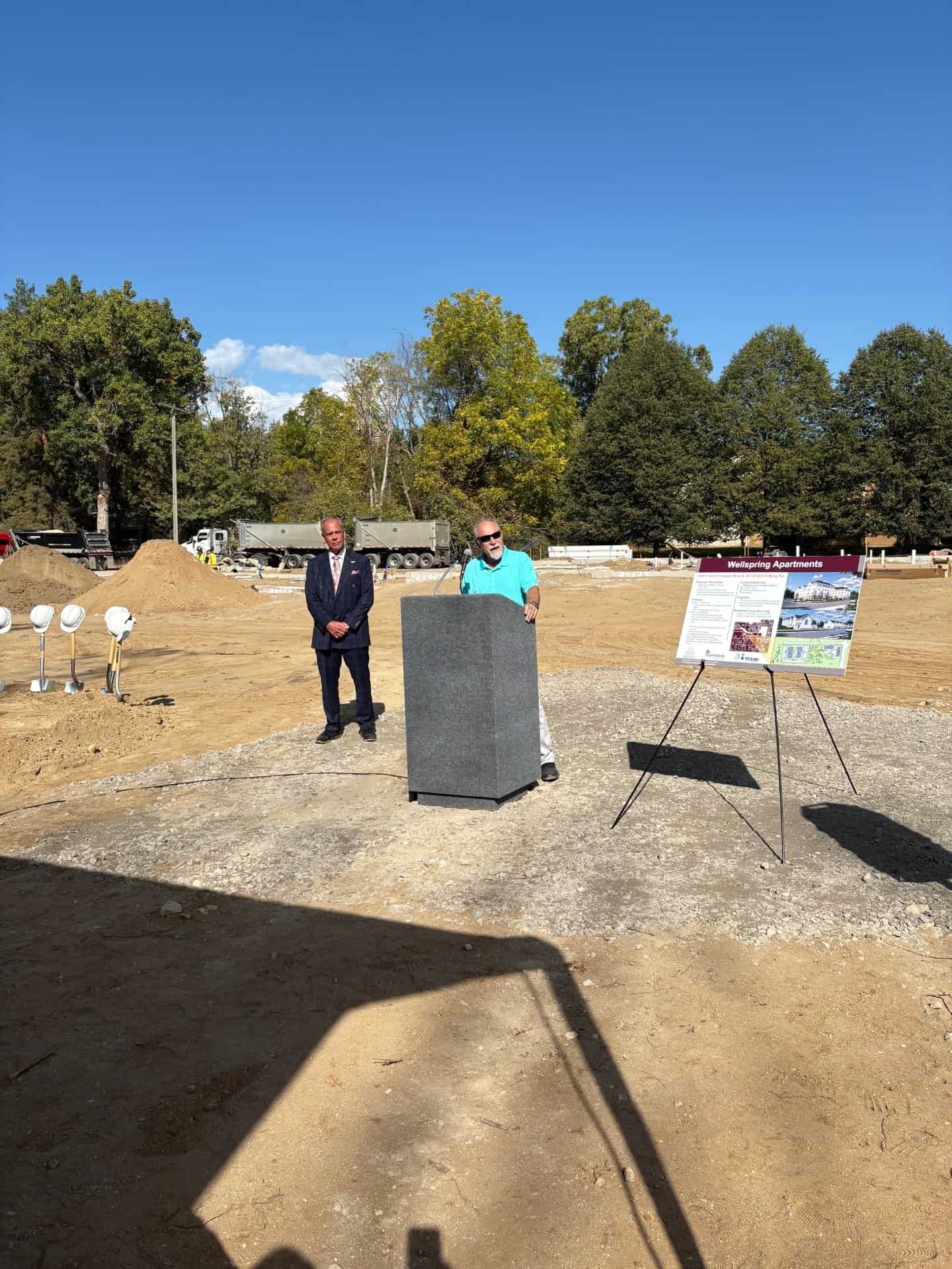 Mike Hirst speaking at podium next to poster and another person standing nearby, at dig site