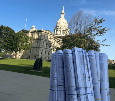 Architectural blueprints in the foreground with the Michigan State Capitol building in the background on a clear day.