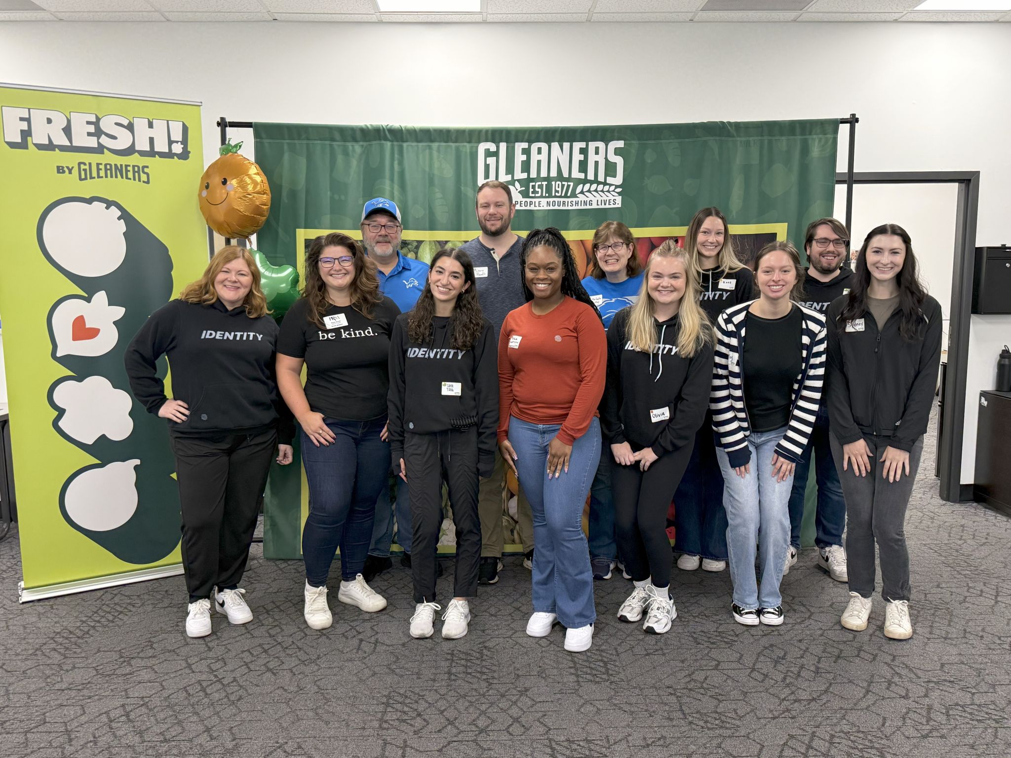 Group of people posing for a photo at a Gleaners event, standing in front of a banner with the Gleaners logo and the text 'FRESH by Gleaners.' Some individuals are wearing shirts with inspirational messages.