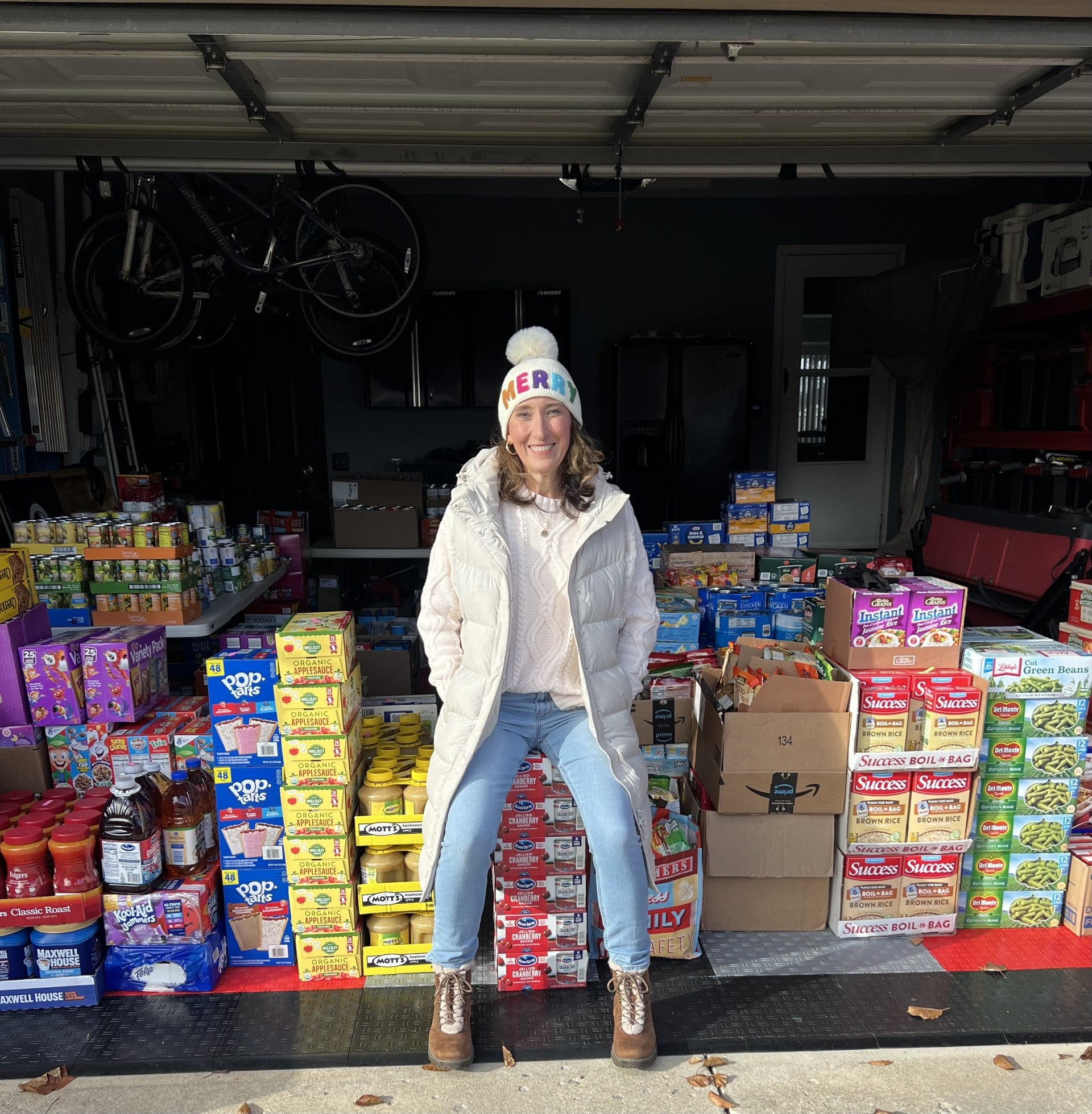 A person stands smiling in a garage filled with boxes and packages of various food items, wearing a winter jacket and a beanie that says 'CHEER.' Bicycles are visible hanging in the background.
