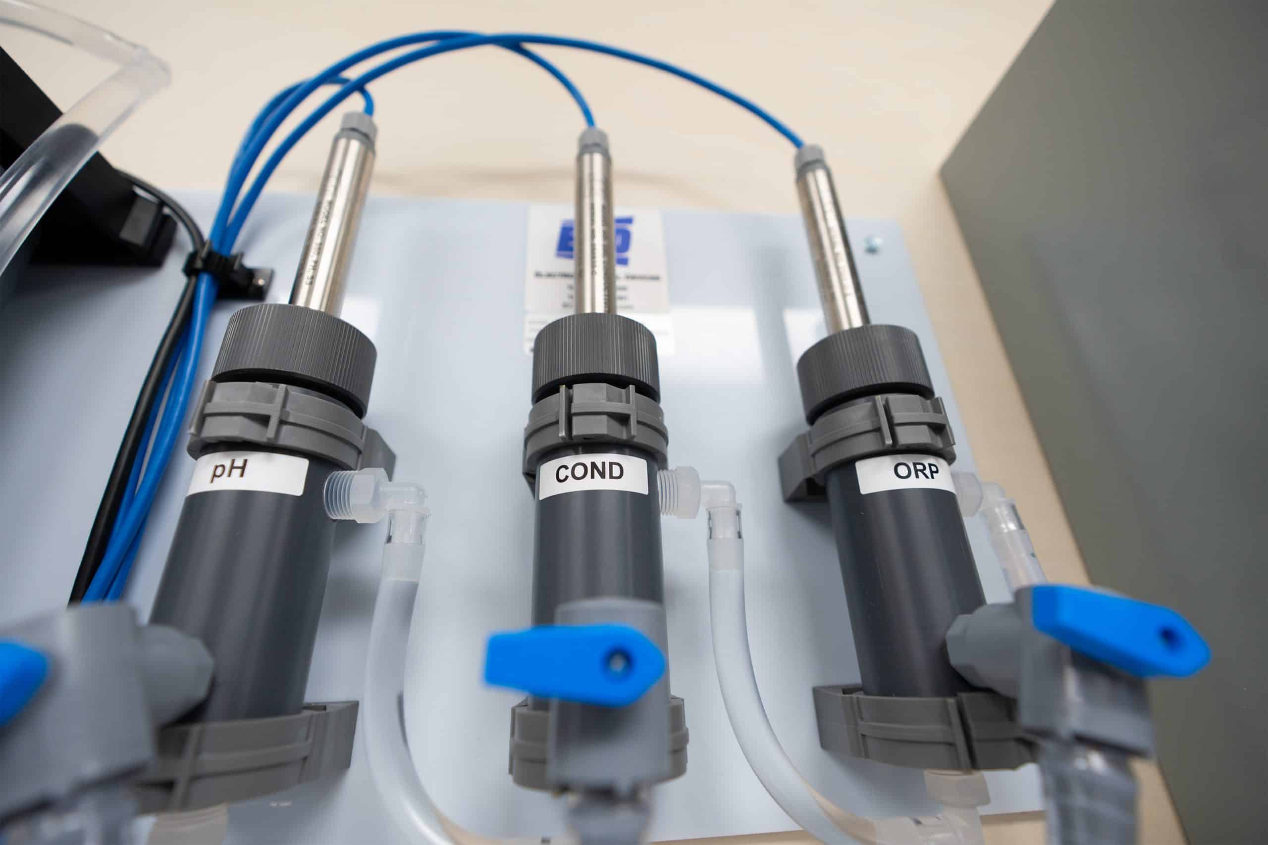Close-up of water monitoring laboratory equipment featuring sensors labeled pH, COND (conductivity), and ORP (oxidation-reduction potential) connected with blue cables.