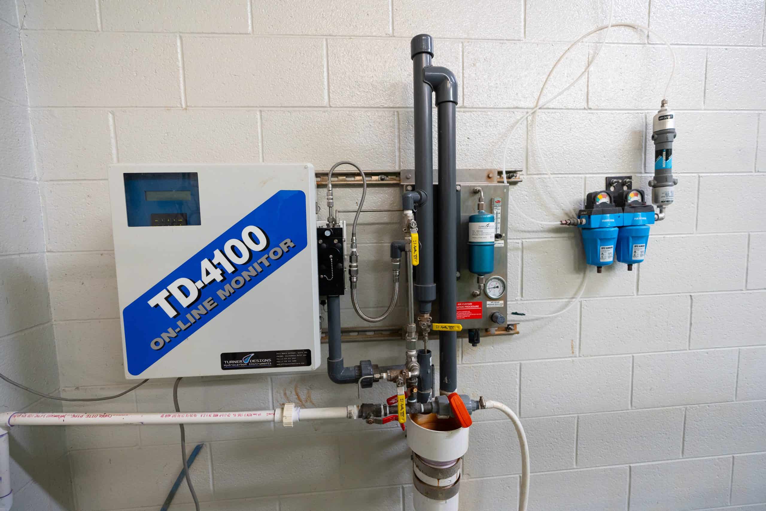TD-4100 oil-in-water monitor mounted on a white cinder block wall, connected to various pipes and valves.